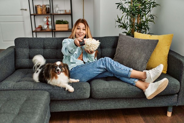 Um sofá espaçoso com um cão, uma tutora, pipoca e um controle remoto.