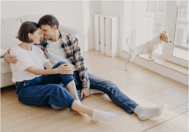 Um casal sentado no chão encostado em um sofá, em um apartamento, com um cachorro pequeno olhando pela janela.